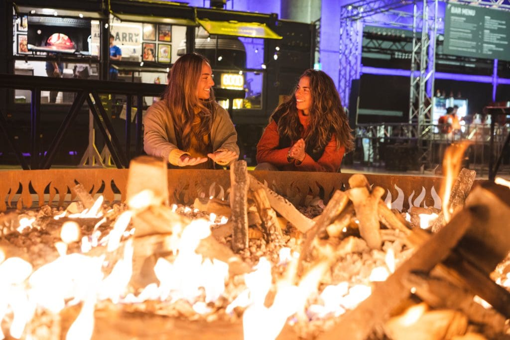 Couple warms up at Vivid Fire Kitchen, Vivid Sydney 2023. Vivid Fire Kitchen brings together Sydney's hottest dining concepts with local and internationally-acclaimed pitmasters and a collection of the finest NSW producers, curated drinks and fire-inspired cocktails for three weeks of flame-fuelled festivities